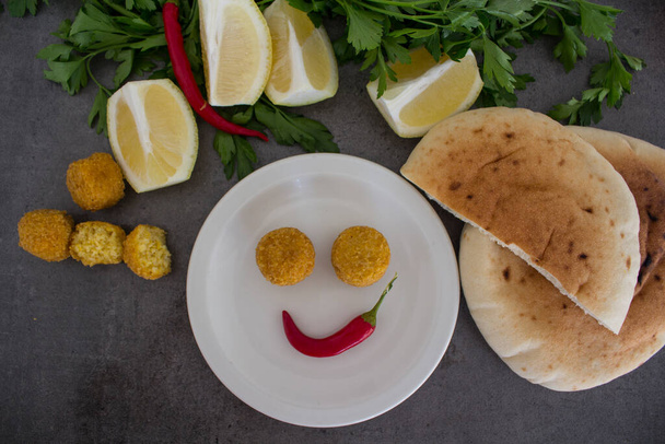 Lächelndes Gesicht aus Essen. Teller mit Falafeln, Fladenbrot, Zitrone und Pfeffer auf dunkelgrauem Tisch. Traditionelles israelisches Essen von oben.  - Foto, Bild