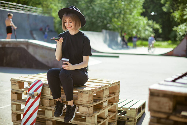 Smiling girl chat on phone sitting on wooden pallet in park with skateboard and cup coffee to go, photo for ad or blog - Fotografie, Obrázek