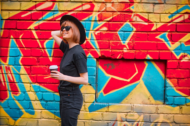 Hezké mladé usmívající se dívka s šálkem kávy v černém klobouku a sluneční brýle stojí proti cihlové zdi s barevnými graffiti - Fotografie, Obrázek