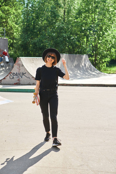Pretty smiling girl carrying skateboard walking in skatepark in sunglasses, hat and black casual clothes - Zdjęcie, obraz