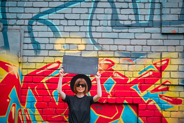 Young girl with cardboard sign of protest above head stand against brick wall with colorful graffiti - Foto, Imagem