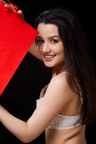 Nuori kaunis nainen valkoisissa alusvaatteissa punaisella paperilla ostoskassi aiheuttaa ja hymyilee. mustalla eristetyllä taustalla. - Valokuva, kuva