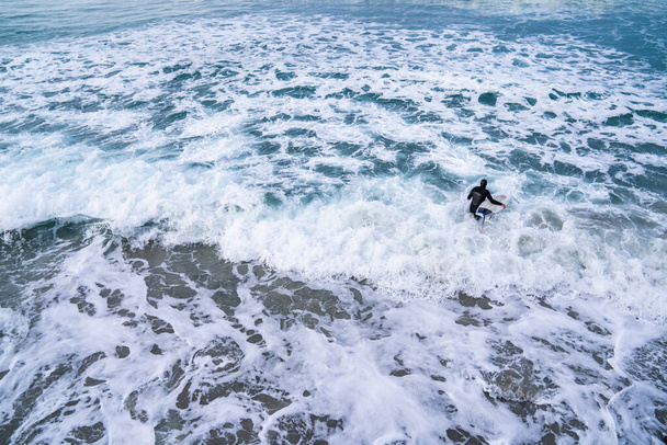 Surfista con traje de neopreno con tabla de surf viendo olas del océano estrellarse sobre rocas en la playa de St. Clair, Dunedin, Nueva Zelanda
. - Foto, imagen
