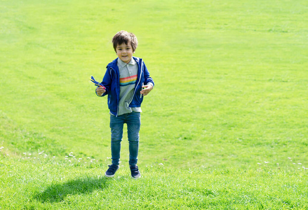 Enfant portrait pleine longueur sautant dans l'herbe verte, mignon petit garçon jouet en plastique avion, enfant s'amusant à jouer en plein air au printemps ou en été champ sur une journée ensoleillée.  - Photo, image