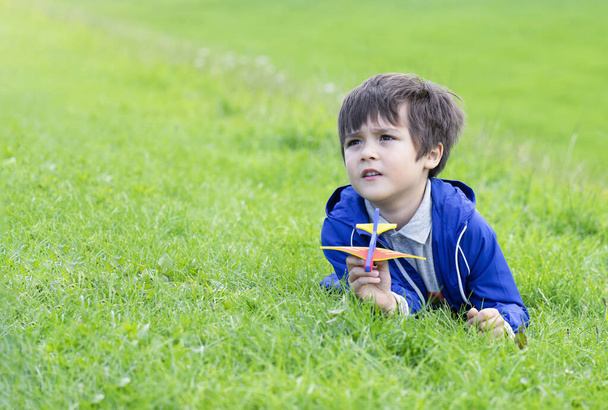 緑の芝生の上に横たわる肖像画の子供の男の子と好奇心の顔を見上げ、緑のフィールドでおもちゃの飛行機で遊ぶアクティブな子供、公園で遊ぶ子供、子供の屋外活動のコンセプト. - 写真・画像