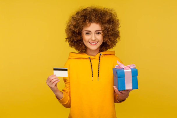 Electronic money and shopping. Portrait of happy curly-haired young woman in urban style hoodie showing credit card and gift box, smiling to camera. indoor studio shot isolated on yellow background - Photo, image