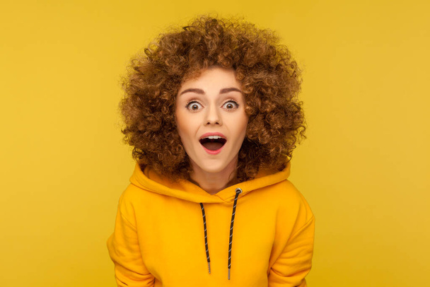 Oh my god, wow! Portrait of surprised curly-haired woman in urban style hoodie looking at camera with mouth open in amazement, expressing shock, astonishment. indoor studio shot, yellow background - Фото, изображение