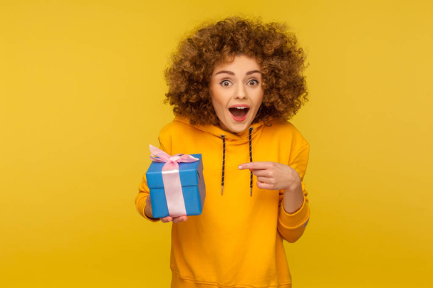 Portrait of funny amazed curly-haired woman in urban style hoodie pointing at blue gift box and looking at camera with surprise, shocked expression. indoor studio shot isolated on yellow background - Photo, image