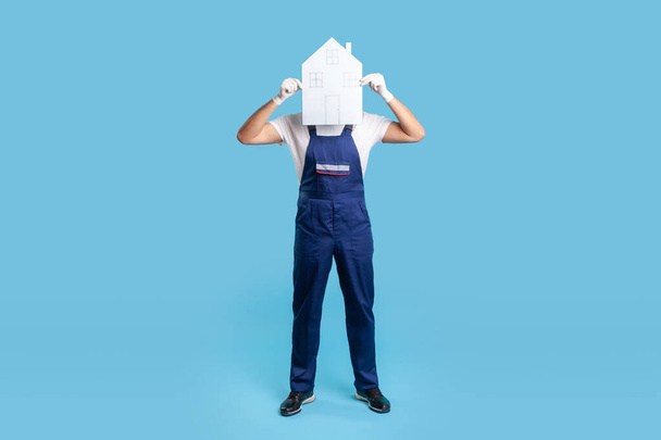 Full body handyman, labourer or mover guy in uniform covering face with paper house. Advertising of home repair service and maintenance, help in relocation. studio shot isolated on blue background - Фото, зображення
