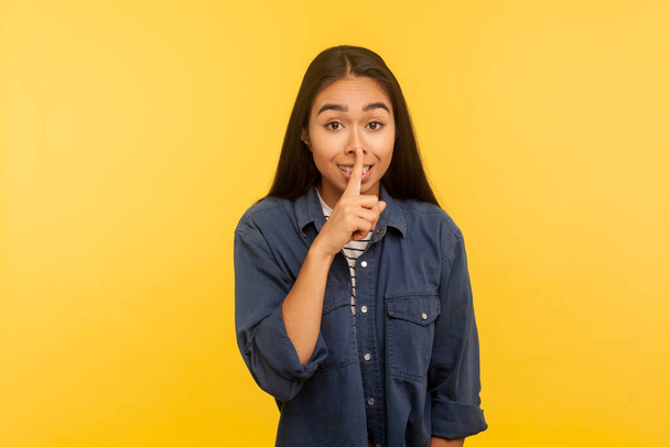 Shh, don't speak! Portrait of happy girl in denim shirt warning of secret, keeping finger on lips asking to stay silent, mystery and confidential information. indoor studio shot, yellow background - Foto, Imagen