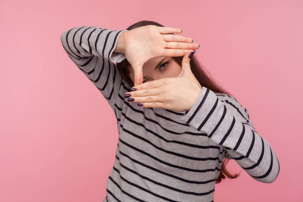 Portrait of woman in striped sweatshirt imitating to take picture, looking through finger frame with attentive view, focusing and cropping image. indoor studio shot isolated on pink background - Photo, Image