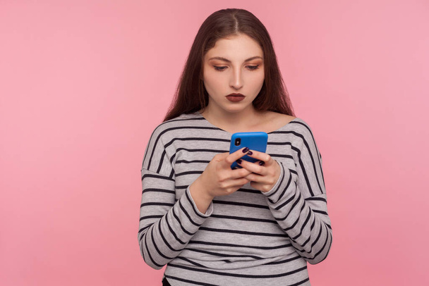Portrait of serious attentive young woman typing on cell phone, reading blog, chatting in social network, using mobile app for online communication. indoor studio shot isolated on pink background - Фото, изображение