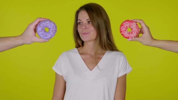 Γυναίκα σε ένα λευκό T-shirt κοιτάζει γύρω και δεν μπορεί να αποφασίσει ποιο ντόνατ να φάει - Πλάνα, βίντεο