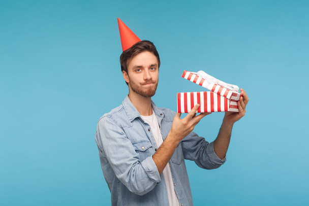 Portrait of frustrated man with party cone hat holding opened gift box and looking at camera with disappointed expression, unpacking present, dissatisfied with awful birthday surprise. studio shot - 写真・画像