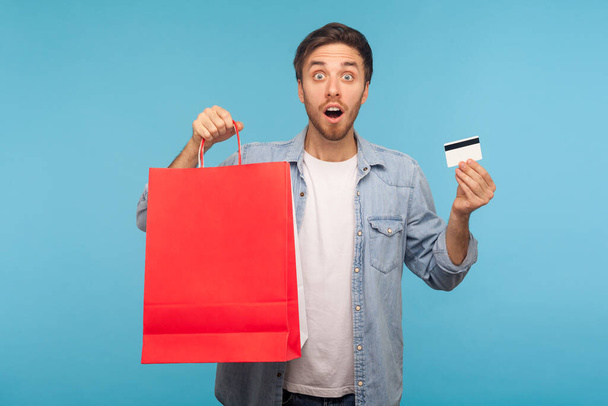 Portrait of amazed handsome man in stylish denim shirt holding credit card and shopping bags with blank space for advertise text, looking with shocked expression. indoor studio shot, blue background - Foto, afbeelding