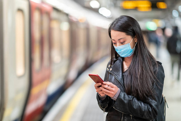 Chinese woman wearing face mask at underground train station and keeping social distance - young asian woman waiting for the train and holding a smartphone - health and travel concepts - Photo, image