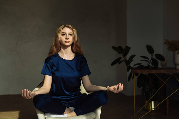 Młoda kobieta medytująca w fotelu w domu. Młode kobiety medytujące ze znakiem mudry na zajęciach jogi lub w domu. Dziewczyna praktyka siedzi na fotelu z lotosami stanowią skrzyżowane nogi. - Zdjęcie, obraz
