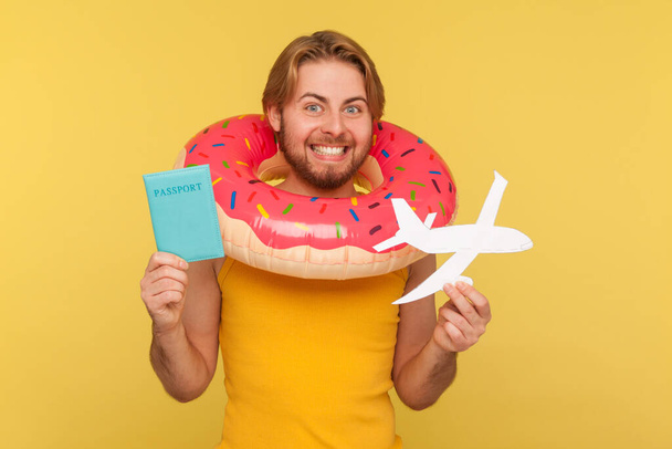 Excited optimistic funny tourist guy in undershirt standing with pink donut rubber ring, holding passport document and airplane mockup, rejoicing travel tour. studio shot isolated on yellow background - Foto, afbeelding