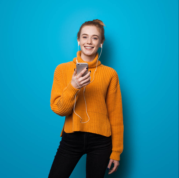 Femme au gingembre caucasien avec des taches de rousseur souriant à la caméra tout en écoutant de la musique et tenant un téléphone sur le mur bleu studio - Photo, image
