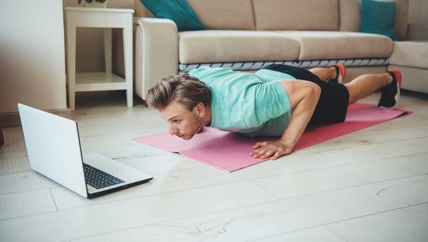 Blonde caucasian man doing push ups at home during the lockdown using a laptop - 写真・画像