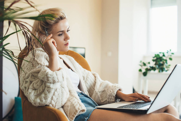 Gedachte blanke vrouw met blond haar met behulp van een laptop thuis met een warme gebreide trui - Foto, afbeelding