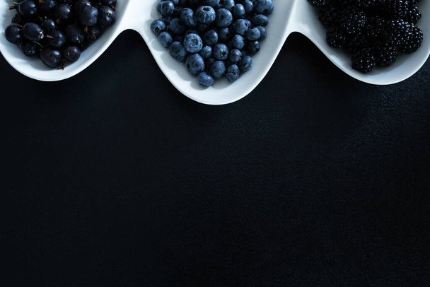 Set of different types of black berries in a white plate on a black table. Stylish seasonal vitamins. - Fotografie, Obrázek