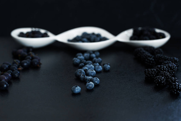 Set of different types of black berries in a white plate on a black table. Stylish seasonal vitamins. - Photo, image