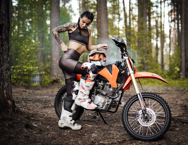 Татуйована дівчина гонщика в мотокросовому одязі з напівголим торсом позує біля свого велосипеда в лісі
 - Фото, зображення