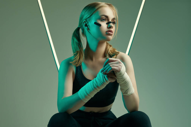 Levendig portret van sterk mooi meisje met blond haar, sportfiguur, zelfverzekerde look en vuisten in beschermende boksen verband poseren op kleurrijke neon lichte achtergrond - Foto, afbeelding
