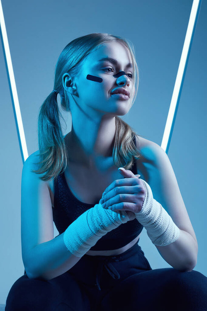 Portrait vif d'une belle fille forte avec des cheveux blonds, une silhouette sportive, un regard confiant et des poings dans des bandages de boxe protecteurs posant sur un fond lumineux néon coloré - Photo, image
