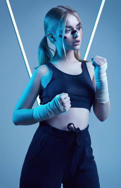 Lebendiges Porträt eines starken, schönen Mädchens mit blonden Haaren, sportlicher Figur, selbstbewusstem Blick und Fäusten in schützenden Boxverbänden, die auf buntem Neonlicht-Hintergrund posieren - Foto, Bild