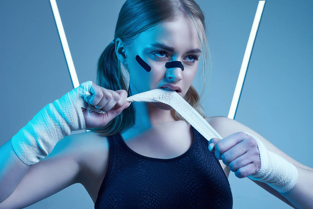 Vivid portrait of strong beautiful girl with blonde hair, sports figure, confident look and fists in protective boxing bandages posing on colorful neon light background - Foto, Imagem