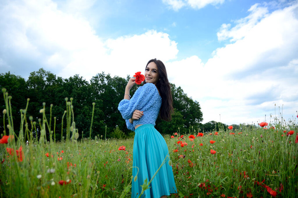 Summer season, young woman in poppy field. Beautiful brunette wearing blue skirt, blue top surrounded by green grass and poppies. - Photo, Image