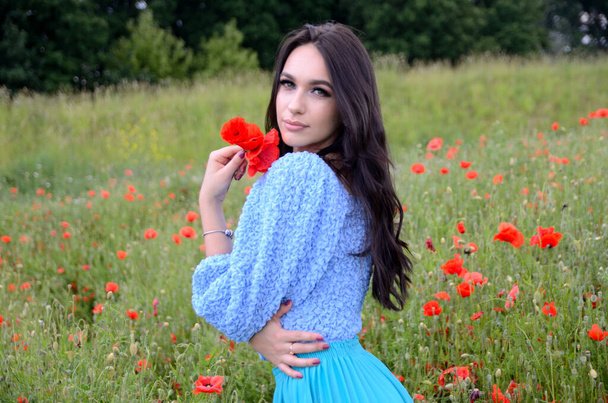 Nyári szezon, fiatal nő a mákmezőn. Gyönyörű barna ruhás kék szoknya, kék felső körül zöld fű és mák. - Fotó, kép