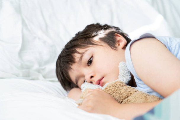  Dzieciak 7-latek leżący na łóżku, śpiące dziecko budzące się rano w swoim pokoju z porannym światłem, słodki chłopczyk śpiący z psią zabawką w łóżku. Opieka zdrowotna nad dziećmi lub problemy ze snem u młodych - Zdjęcie, obraz