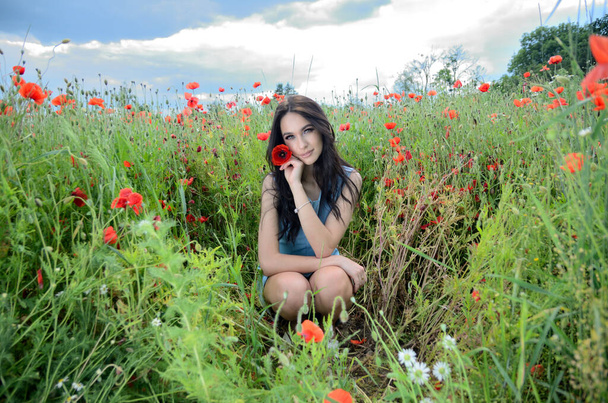 Nyári szezon, fiatal nő a mákmezőn. Gyönyörű barna ruhás kék ruha, körülvéve zöld fű és mák. - Fotó, kép