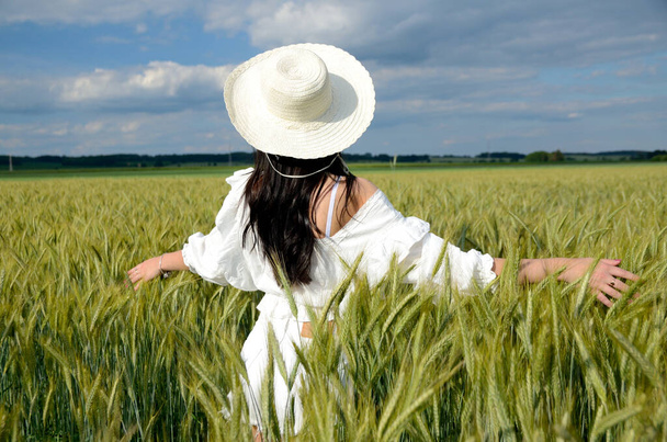 Country girl with white hat surrounded by barley field. Beautiful brunette wearing white top and white shorts. Photo from behind, without face. - Fotoğraf, Görsel