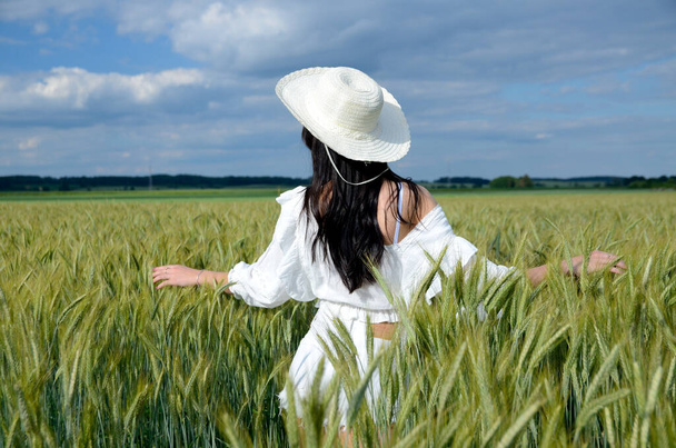 大麦のフィールドに囲まれた白い帽子を持つ国の女の子。白の上と白のショートパンツを着た美しいブルネット。後ろから顔のない写真. - 写真・画像
