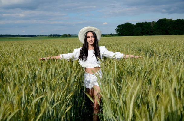 大麦のフィールドに囲まれた白い帽子を持つ国の女の子。美しいブルネットは白い上と白いショートパンツを着て. - 写真・画像