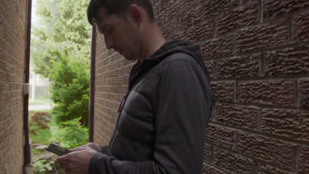 High quality 4k video of a young man who is texting messages on his cellphone standing in a shade of the red bricks wall. Guy hiding between the walls and holding mobile phone in his hands. - Metraje, vídeo