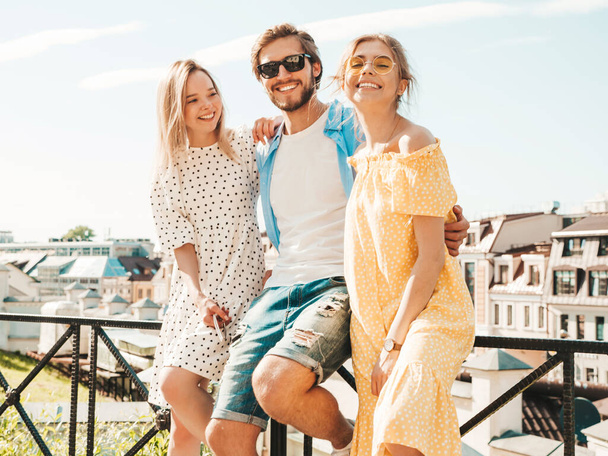 Group of young three stylish friends posing in the street. Fashion man and two cute girls dressed in casual summer clothes. Smiling models having fun in sunglasses.Cheerful women and guy outdoors - Fotografie, Obrázek