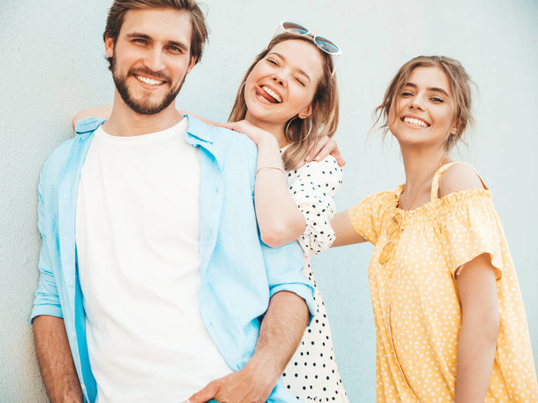 Group of young three stylish friends posing in the street. Fashion man and two cute girls dressed in casual summer clothes. Smiling models having fun near wall.Cheerful women and guy outdoors - Photo, image