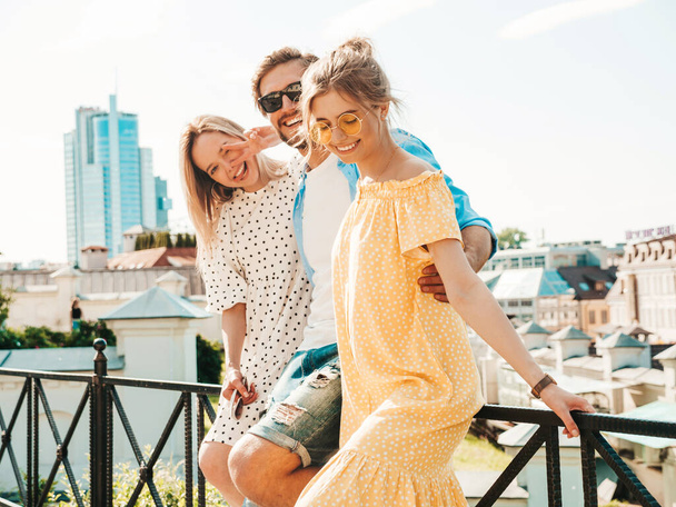 Group of young three stylish friends posing in the street. Fashion man and two cute girls dressed in casual summer clothes. Smiling models having fun in sunglasses.Cheerful women and guy outdoors - Zdjęcie, obraz