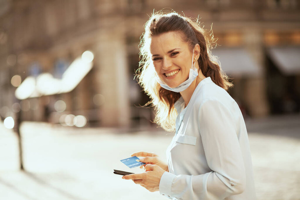 Life during covid-19 pandemic. Portrait of happy young woman in blue blouse with credit card and medical mask buying online outdoors on the city street. - Fotoğraf, Görsel