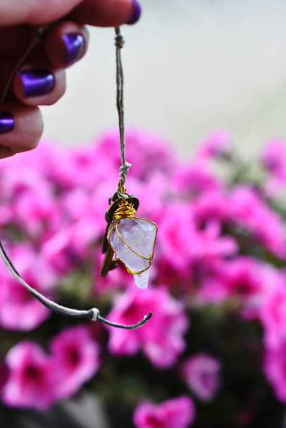 Nahaufnahme eines hausgemachten Amethyst-Pendels gegen leuchtend rosa Petunienblüten.  - Foto, Bild