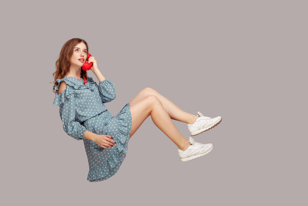 Charming girl in ruffle dress holding phone handset levitating, hovering floating air and having conversation on telephone, looking up with dreamy expression. studio shot isolated gray background, - Foto, Bild