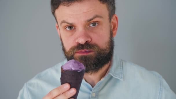 Κοντινό πλάνο ενός γενειοφόρου άντρα που τρώει μωβ παγωτό σε μαύρο κύπελλο βάφλας σε γκρι φόντο τοίχου - Πλάνα, βίντεο