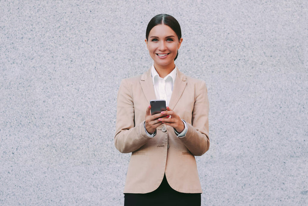Portrait young attractive successful business woman in suit use smart phone and smile gray background. Beautiful happy female entrepreneur hold mobile phone in hands. Millennial girl corporate lawyer - Φωτογραφία, εικόνα