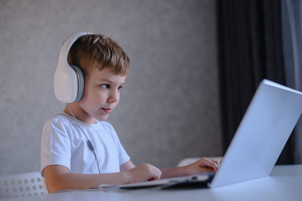 Cute diligent boy is typing on the keyboard and is carefully looking at the laptop screen. Caucasian schoolboy sits at a table, listens to music on headphones and does homework. Distance education. - Foto, immagini