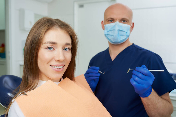 Dentist in medical face mask is holding a dental mirror and dental explorer near a smiling female patient in a dental chair. Doctor with a patient who is demonstrating a result of the tooth treatment - Zdjęcie, obraz
